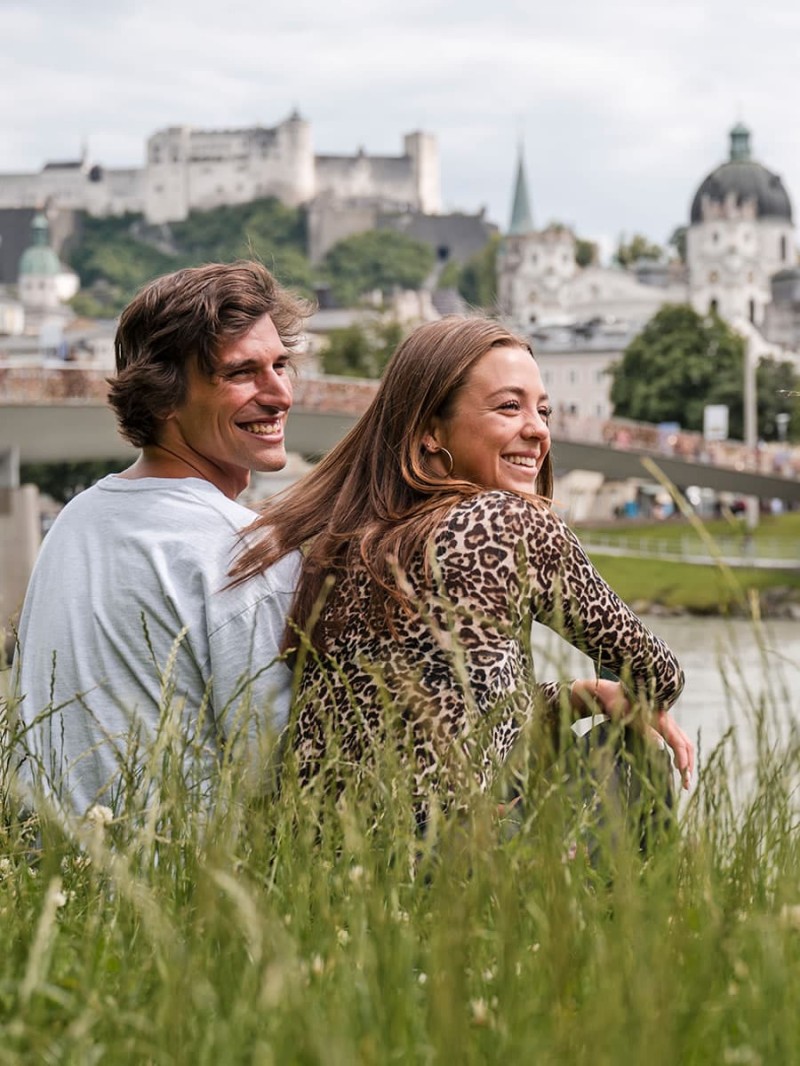 Vacationing couple sitting on the Salzach riverbank overlooking the Old Town